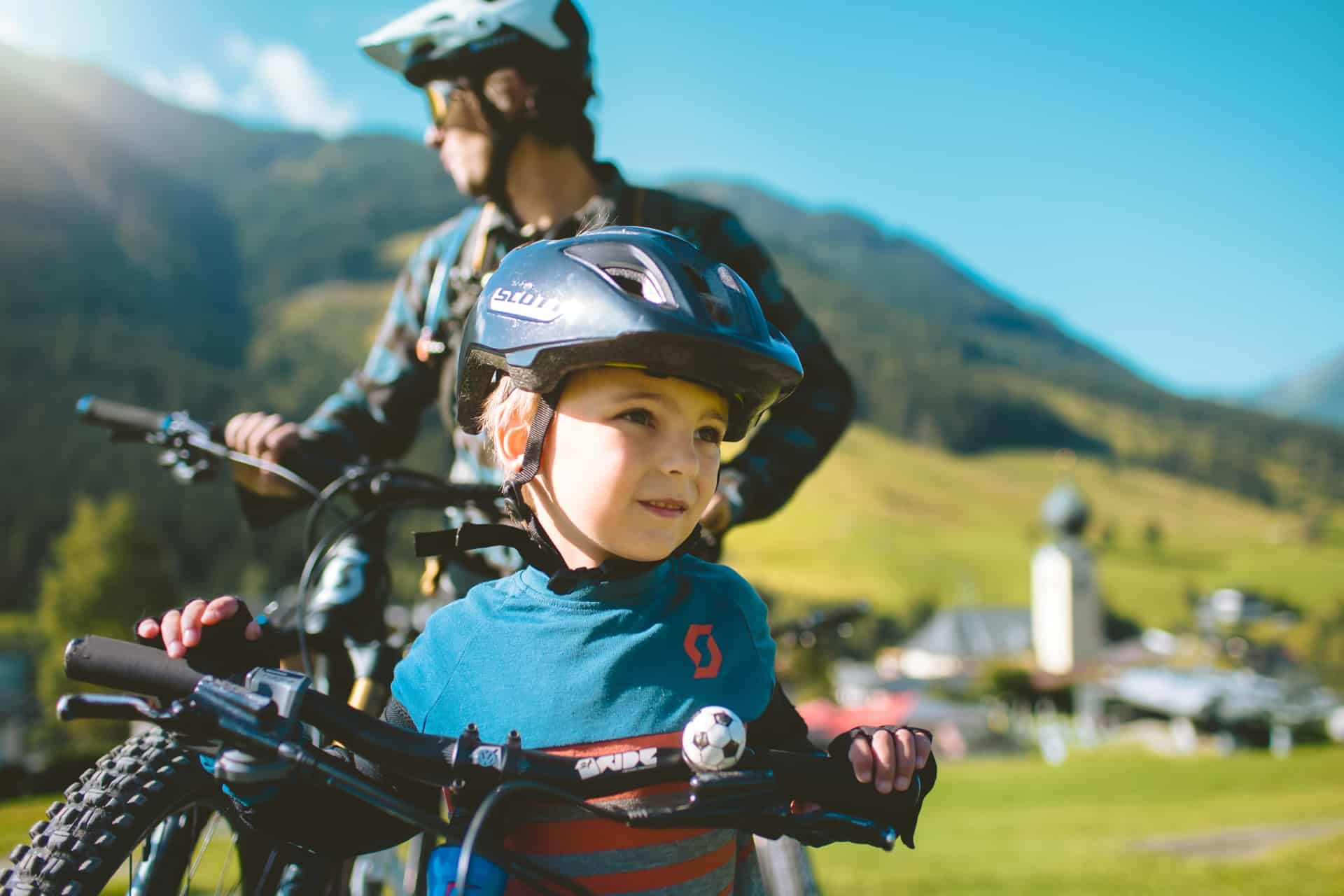 Kind im Bikecircus. Im Hintergrund steht ein Mann und blickt in die Ferne. Noch weiter im Hintergrund ist eine grüne Landschaft zu sehen