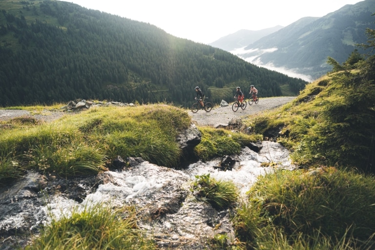 3 Biker auf Bikestrecke, die sich neben einem Gebirgsbach entlang zieht